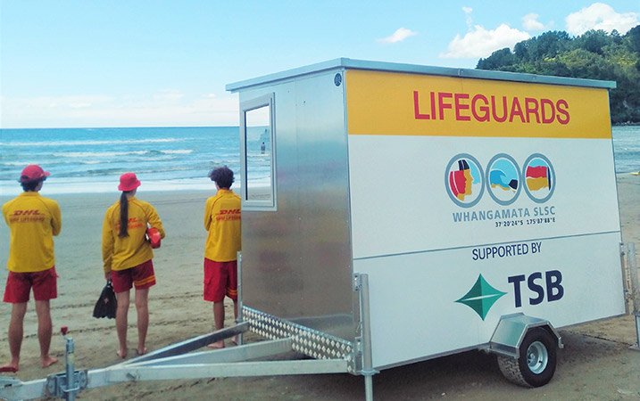 Three lifeguards at the beach standing next to the Whangamata SLSC trailer