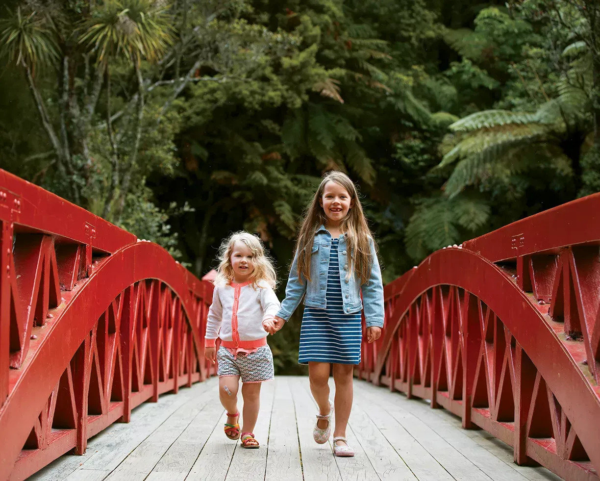 Two children walking on a red bridge surrounded by greenery